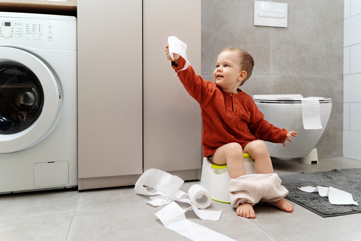 A toddler sitting on a potty and holding a roll of toilet paper, with Step By Step Nursery Dubai supporting toilet independence through nurturing guidance.