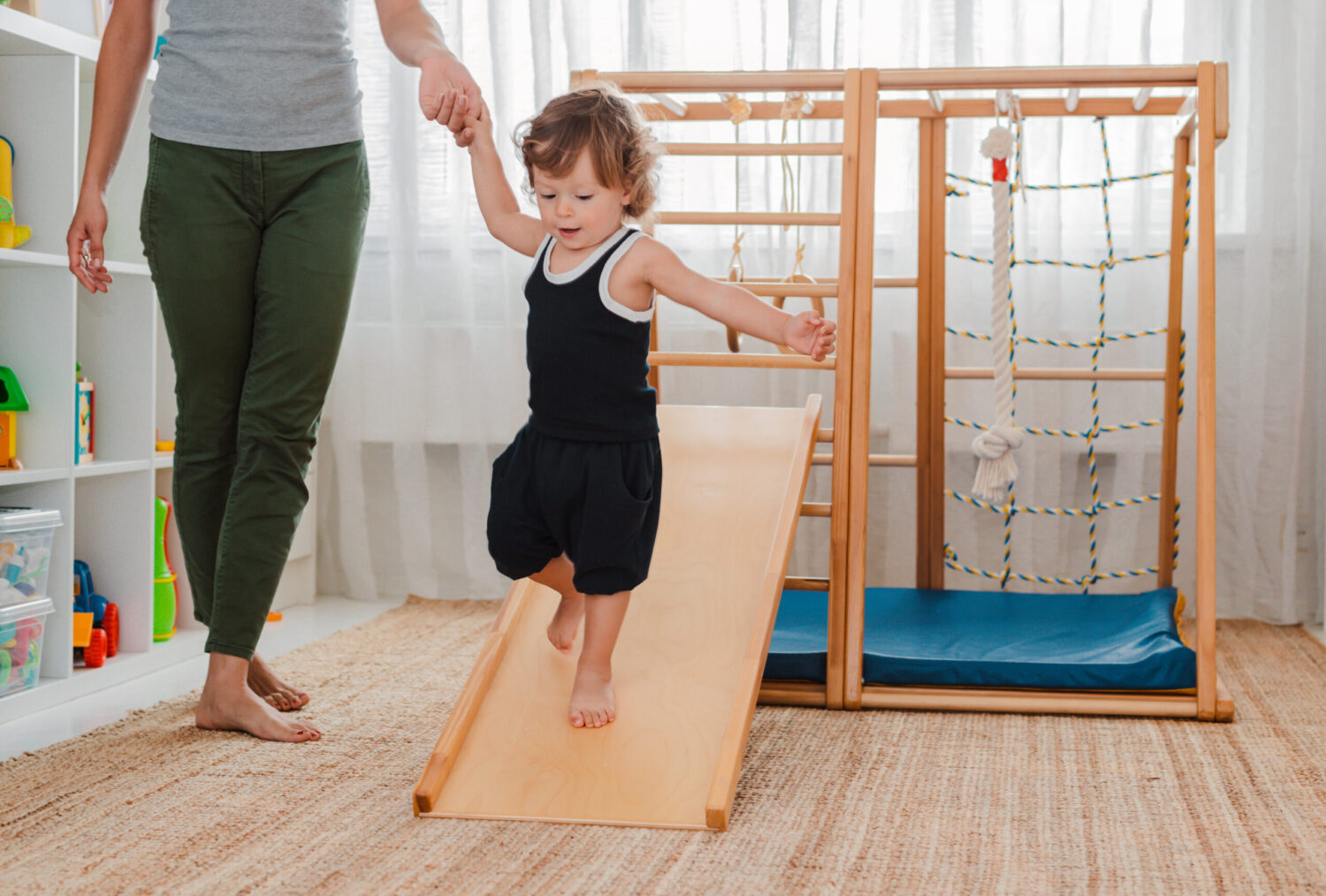 Teacher holds toddler's hand as they walk down a slide, practicing balance and coordination