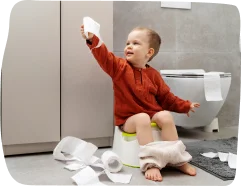 A toddler sitting on a potty and holding a roll of toilet paper, with Step By Step Nursery Dubai supporting toilet independence through nurturing guidance.